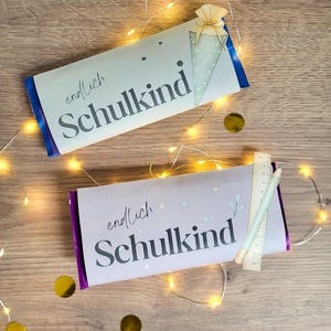 Puede incluir: Dos barras de chocolate rectangulares envueltas en papel con el texto "endlich Schulkind" impreso en negro. La barra superior presenta una ilustración de un cono verde, mientras que la inferior incluye una regla de madera de 12 cm y un lápiz.