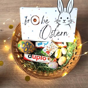 May include: A wicker basket filled with Easter treats, including chocolate bars, decorated eggs, and a box with a bunny illustration and the words "Frohe Ostern". The basket is decorated with fairy lights and gold confetti.