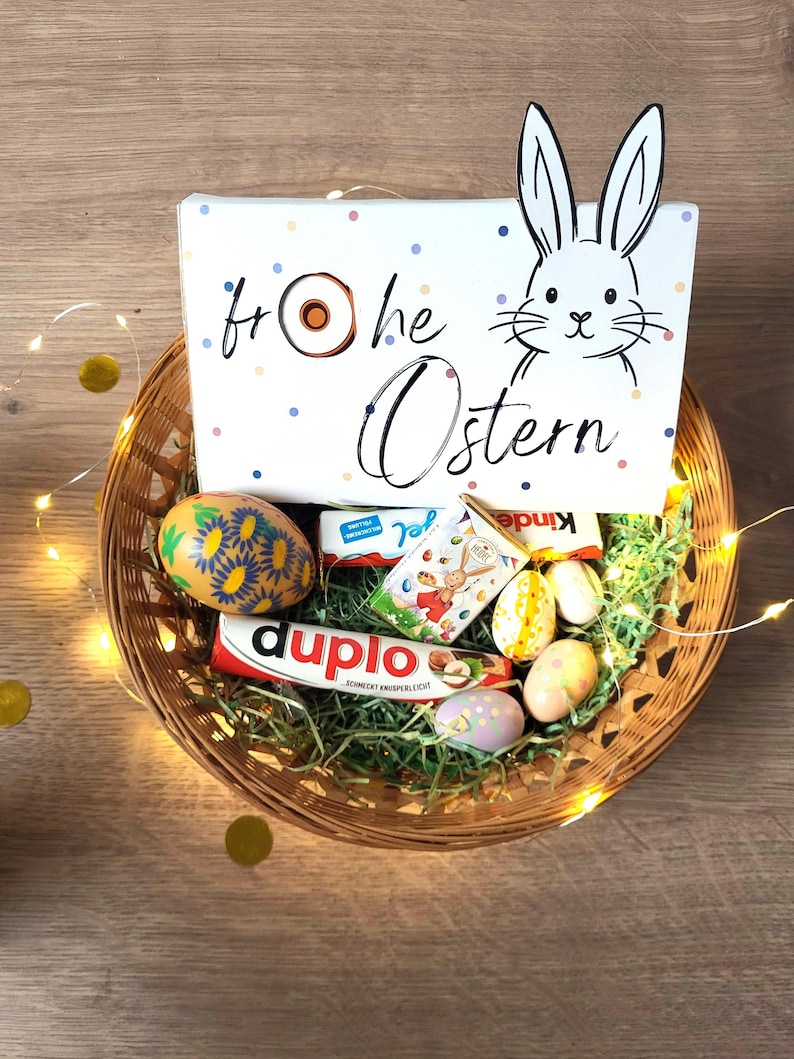 May include: A wicker basket filled with Easter treats, including chocolate bars, decorated eggs, and a box with a bunny illustration and the words "Frohe Ostern". The basket is decorated with fairy lights and gold confetti.
