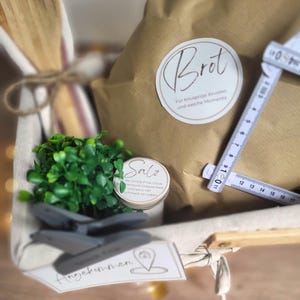 May include: A gift basket containing a brown paper bag labeled 'Brot', a small jar labeled 'Salz', a wooden spoon, and a green plant. A white folding ruler is placed across the bag, showing measurements up to 17 inches. A tag labeled 'Angekommen' is attached to the front.