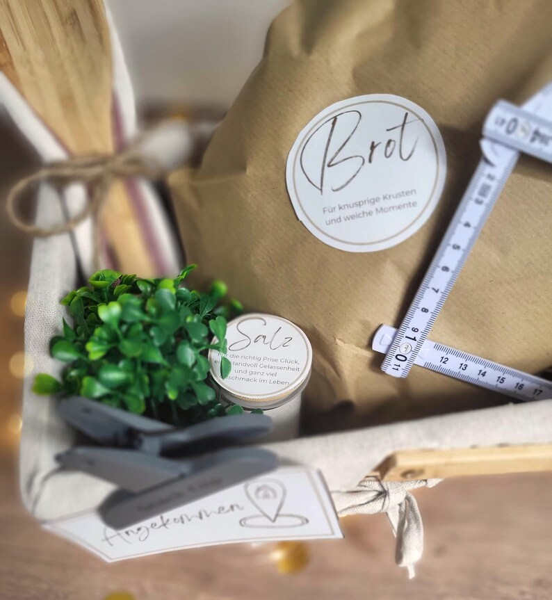 May include: A gift basket containing a brown paper bag labeled 'Brot', a small jar labeled 'Salz', a wooden spoon, and a green plant. A white folding ruler is placed across the bag, showing measurements up to 17 inches. A tag labeled 'Angekommen' is attached to the front.