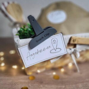 May include: A dark gray plastic snack clip holding a rectangular white card with a gold border. The card features the handwritten text "Angekommen" and a line-art icon of a house inside a location pin symbol, attached to a fabric-lined basket.