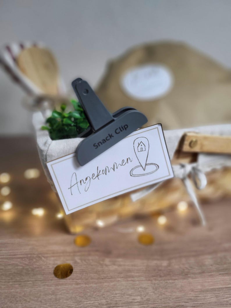 May include: A dark gray plastic snack clip holding a rectangular white card with a gold border. The card features the handwritten text "Angekommen" and a line-art icon of a house inside a location pin symbol, attached to a fabric-lined basket.