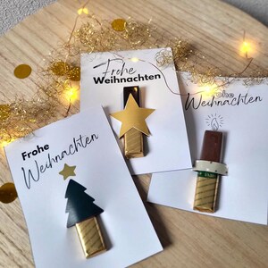 May include: Three white cards with the text "Frohe Weihnachten" and festive chocolate decorations. One card features a gold star, another a Christmas tree, and the last a candle. Gold confetti and string lights are in the background.