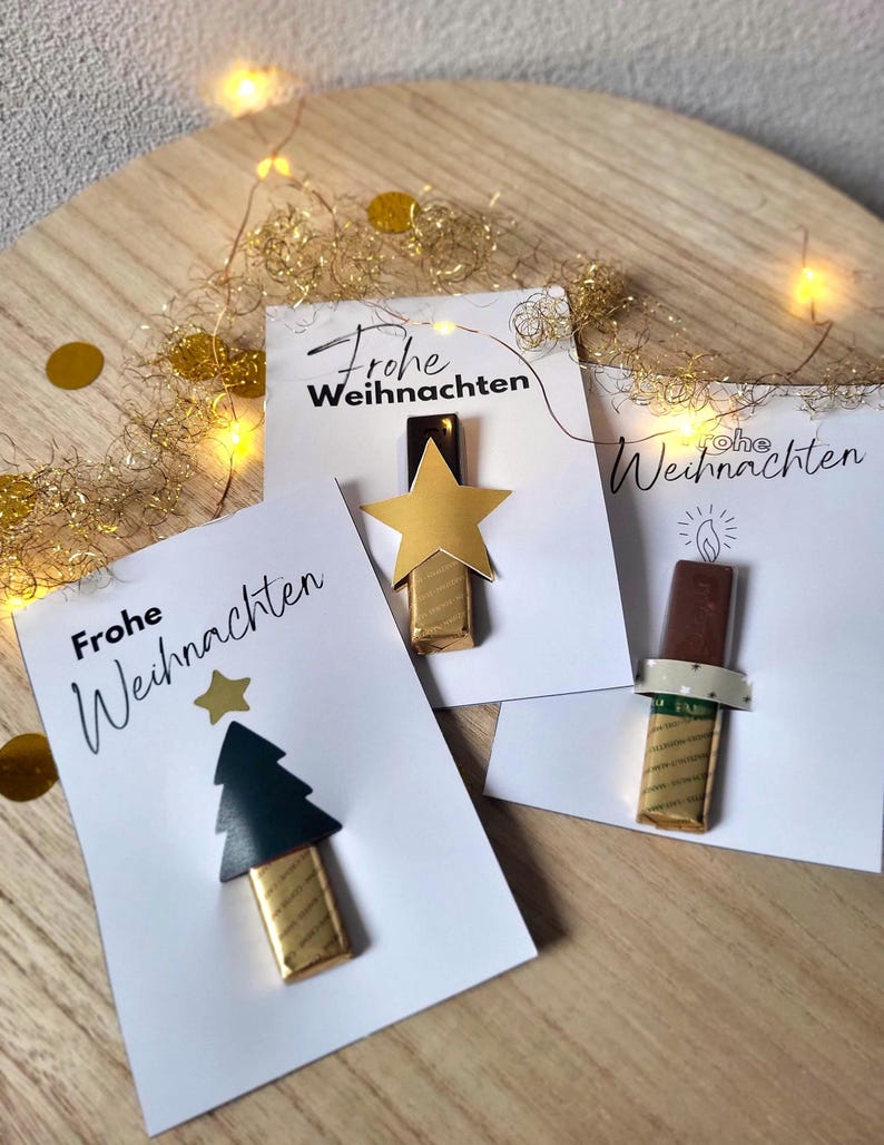 May include: Three white cards with the text "Frohe Weihnachten" and festive chocolate decorations. One card features a gold star, another a Christmas tree, and the last a candle. Gold confetti and string lights are in the background.