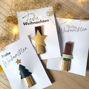 May include: Three white cards with the German phrase "Frohe Weihnachten" printed in black. Each card features a chocolate bar decorated with a gold star, a Christmas tree, and a candle. Gold confetti and string lights are scattered around.