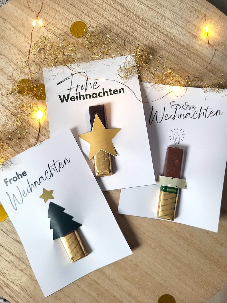 May include: Three white cards with the German phrase "Frohe Weihnachten" printed in black. Each card features a chocolate bar decorated with a gold star, a Christmas tree, and a candle. Gold confetti and string lights are scattered around.