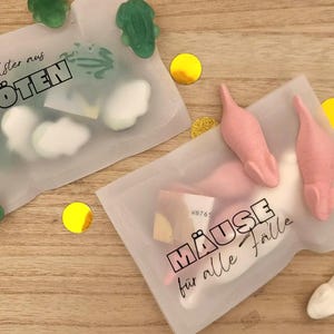 以下が含まれることがあります： 2つの透明なプラスチックバッグ。1つは緑色のカエル型グミとテキスト「ein Polster aus KR&Ouml;TEN」、もう1つはピンクと白のネズミ型キャンディーとテキスト「M&Auml;USE f&uuml;r alle F&auml;lle」が入っています。金色の紙吹雪が散らばっています。