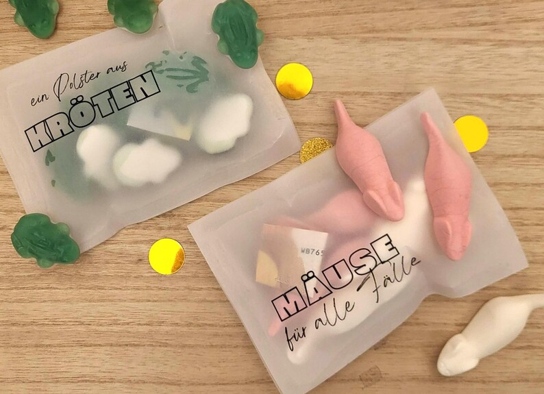 以下が含まれることがあります： 2つの透明なプラスチックバッグ。1つは緑色のカエル型グミとテキスト「ein Polster aus KR&Ouml;TEN」、もう1つはピンクと白のネズミ型キャンディーとテキスト「M&Auml;USE f&uuml;r alle F&auml;lle」が入っています。金色の紙吹雪が散らばっています。