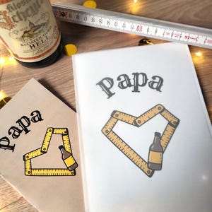 May include: Two cards with the word "Papa" in a decorative font. One card is tan, the other white. Each card features a yellow folding ruler forming a heart shape around a beer bottle illustration. A beer bottle and a ruler are also visible.