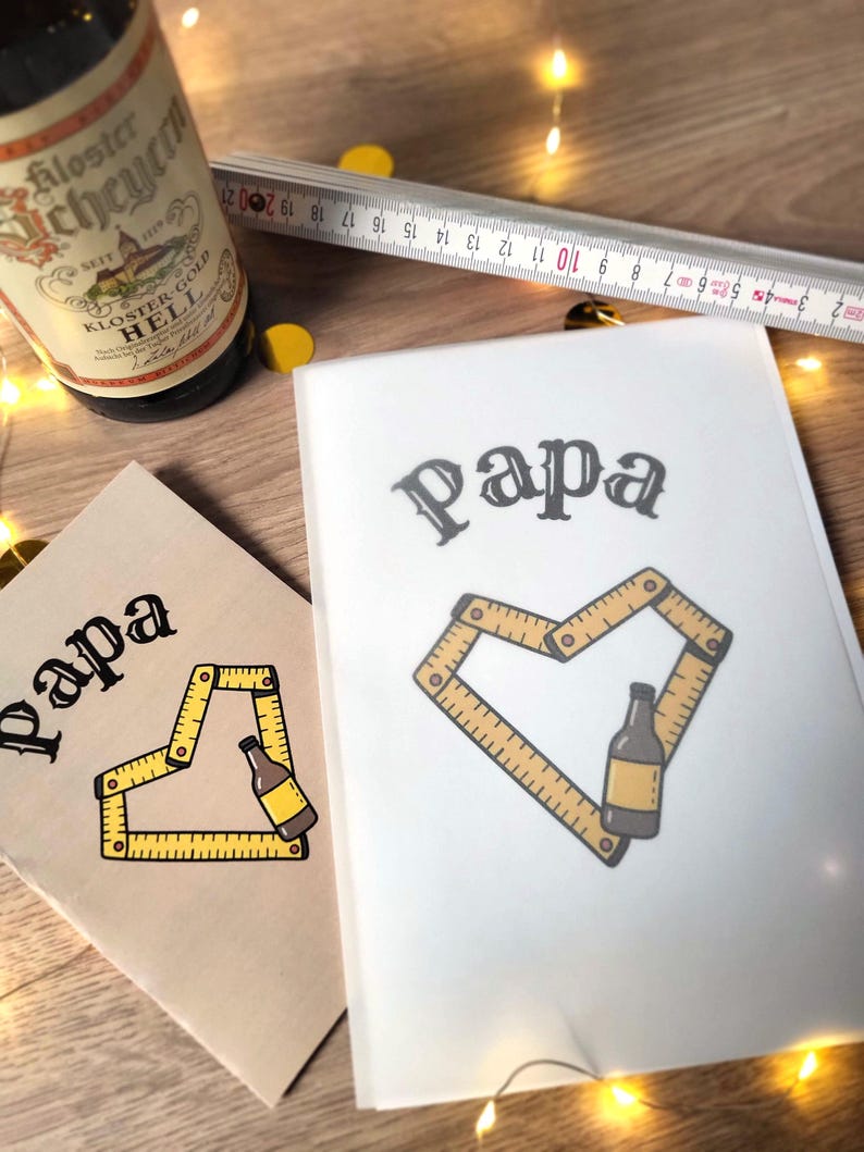 May include: Two cards with the word "Papa" in a decorative font. One card is tan, the other white. Each card features a yellow folding ruler forming a heart shape around a beer bottle illustration. A beer bottle and a ruler are also visible.