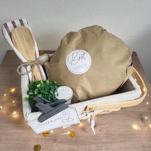 May include: A rectangular woven basket with a fabric liner containing a brown paper-wrapped loaf labeled Brot, a wooden spoon tied with twine to a striped cloth, a small green plant, a jar labeled Salz, a grey snack clip, and a tag labeled Angekommen.