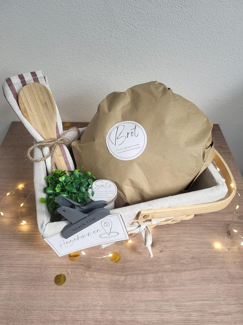 May include: A rectangular woven basket with a fabric liner containing a brown paper-wrapped loaf labeled Brot, a wooden spoon tied with twine to a striped cloth, a small green plant, a jar labeled Salz, a grey snack clip, and a tag labeled Angekommen.