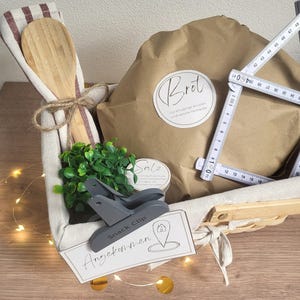 May include: A gift basket containing a wooden spoon, a brown paper-wrapped item labeled Brot, a small green plant, a container labeled Salz, a gray snack clip, and a white card labeled Angekommen, with a folding ruler showing 18.5 inches.