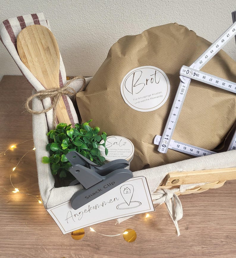 May include: A gift basket containing a wooden spoon, a brown paper-wrapped item labeled Brot, a small green plant, a container labeled Salz, a gray snack clip, and a white card labeled Angekommen, with a folding ruler showing 18.5 inches.
