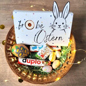 May include: A woven basket filled with Easter treats, including chocolate bars and decorated eggs. A white box with a bunny illustration and the words "Frohe Ostern" is in the basket. The basket is decorated with fairy lights.