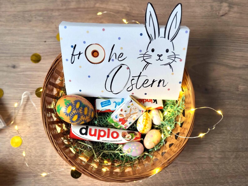 May include: A woven basket filled with Easter treats, including chocolate bars and decorated eggs. A white box with a bunny illustration and the words "Frohe Ostern" is in the basket. The basket is decorated with fairy lights.