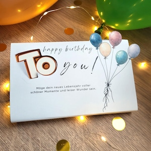 Peut inclure: Une carte de joyeux anniversaire blanche avec l'inscription "To you!" dans une police fantaisiste, accompagnée d'un bouquet de ballons aquarelle. La carte est décorée de confettis dorés et de guirlandes lumineuses, avec des ballons orange et verts en arrière-plan.