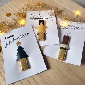 May include: Three white cards with the text "Frohe Weihnachten" and decorative elements. One card features a gold star, another a green tree, and the third a candle. Each card has a chocolate bar attached.