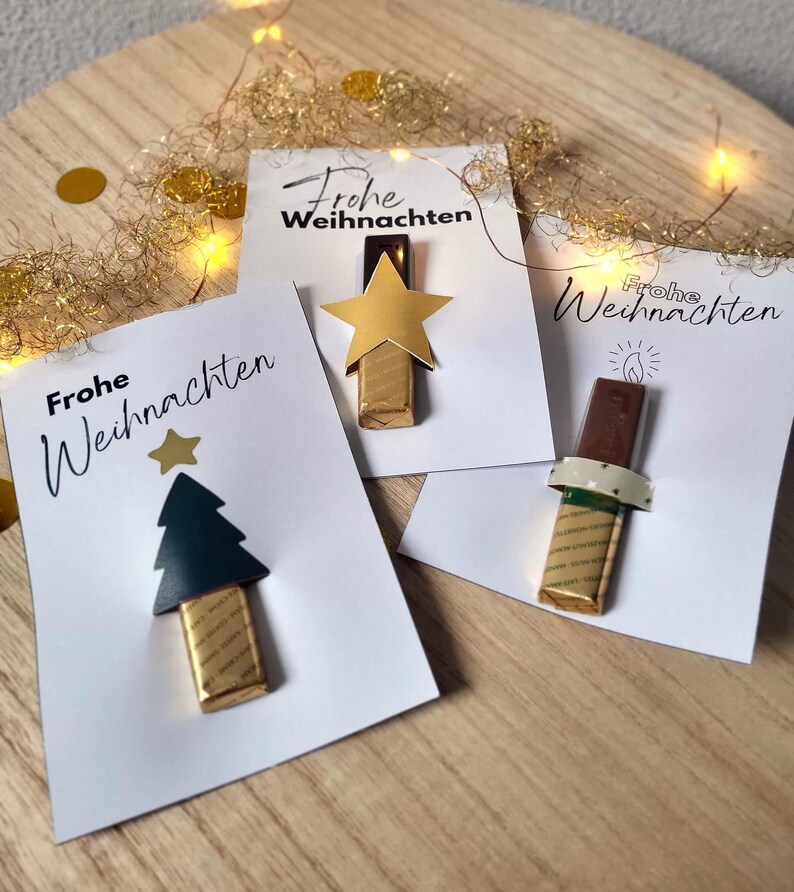 May include: Three white cards with the text "Frohe Weihnachten" and decorative elements. One card features a gold star, another a green tree, and the third a candle. Each card has a chocolate bar attached.