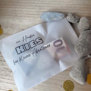 May include: A small white translucent bag with the German text "ein Haufen KIES f&uuml;r kleine Abenteuer" and a ten-euro note inside. Several grey stones and gold confetti surround the bag on a wooden surface.