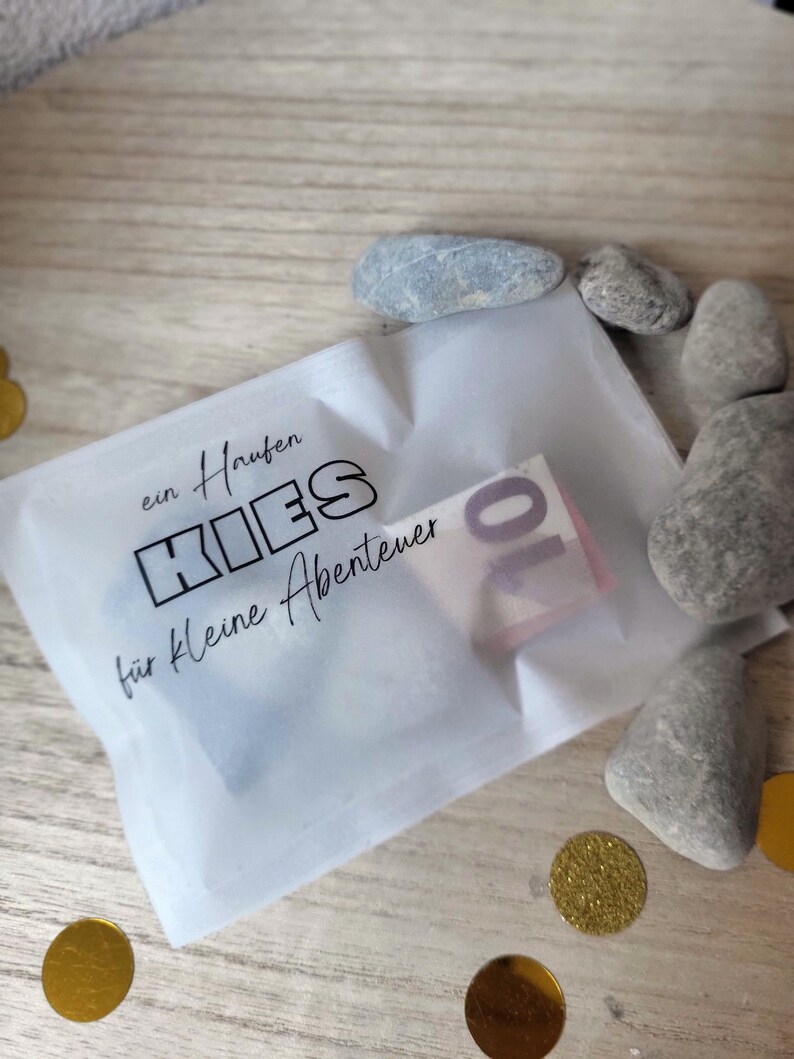 May include: A small white translucent bag with the German text "ein Haufen KIES f&uuml;r kleine Abenteuer" and a ten-euro note inside. Several grey stones and gold confetti surround the bag on a wooden surface.