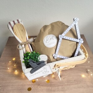 May include: A gift basket containing a brown paper-wrapped package labeled "Brot", a wooden spoon, a small green plant, a gray snack clip, and a white folding ruler measuring up to 50 cm. A tag reads "Angekommen" with a house icon.