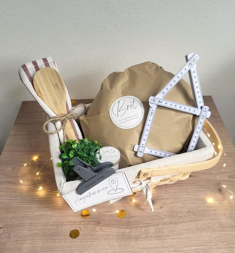 May include: A gift basket containing a brown paper-wrapped package labeled "Brot", a wooden spoon, a small green plant, a gray snack clip, and a white folding ruler measuring up to 50 cm. A tag reads "Angekommen" with a house icon.
