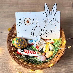 Puede incluir: Una cesta de mimbre llena de golosinas de Pascua, incluyendo huevos de chocolate y barritas de caramelo. Una tarjeta blanca con una ilustración de conejito y las palabras "Frohe Ostern" está en la cesta. La cesta está decorada con hierba de papel verde y luces de cadena.