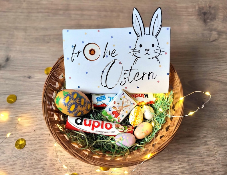 May include: A woven basket filled with Easter treats, including chocolate eggs and candy bars. A white card with a bunny illustration and the words "Frohe Ostern" is in the basket. The basket is decorated with green paper grass and string lights.
