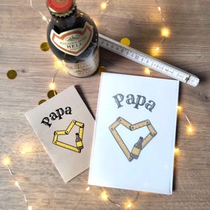 May include: Two greeting cards with the word "Papa" and a heart design made of a ruler and a bottle. A bottle of beer and a folding ruler are also present. The image is decorated with string lights and gold confetti.