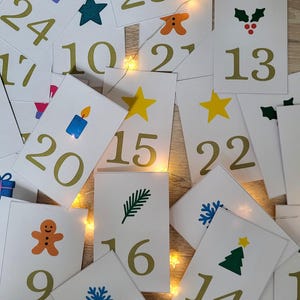 Puede incluir: Un conjunto de tarjetas rectangulares blancas con números dorados e ilustraciones festivas. Las tarjetas presentan imágenes de un guante, estrella, vela, hombre de jengibre, copo de nieve y árbol de Navidad. Las tarjetas están esparcidas sobre una superficie de madera.