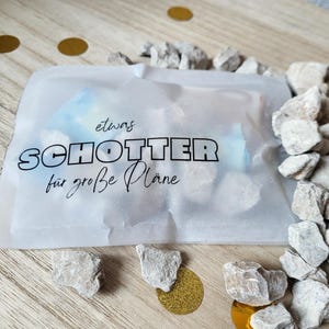 May include: A cellophane bag with German text, surrounded by small, light-coloured rocks and gold confetti on a wooden surface. The text reads "etwas SCHOTTER f&uuml;r gro&szlig;e Pl&auml;ne".