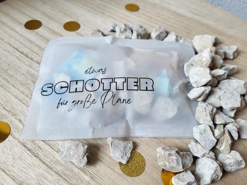May include: A cellophane bag with German text, surrounded by small, light-coloured rocks and gold confetti on a wooden surface. The text reads "etwas SCHOTTER f&uuml;r gro&szlig;e Pl&auml;ne".