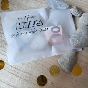 May include: A white translucent paper envelope with the text "ein Haufen KIES f&uuml;r kleine Abenteuer" and a ten-euro note inside. The envelope is surrounded by small grey stones and gold confetti on a wooden surface.