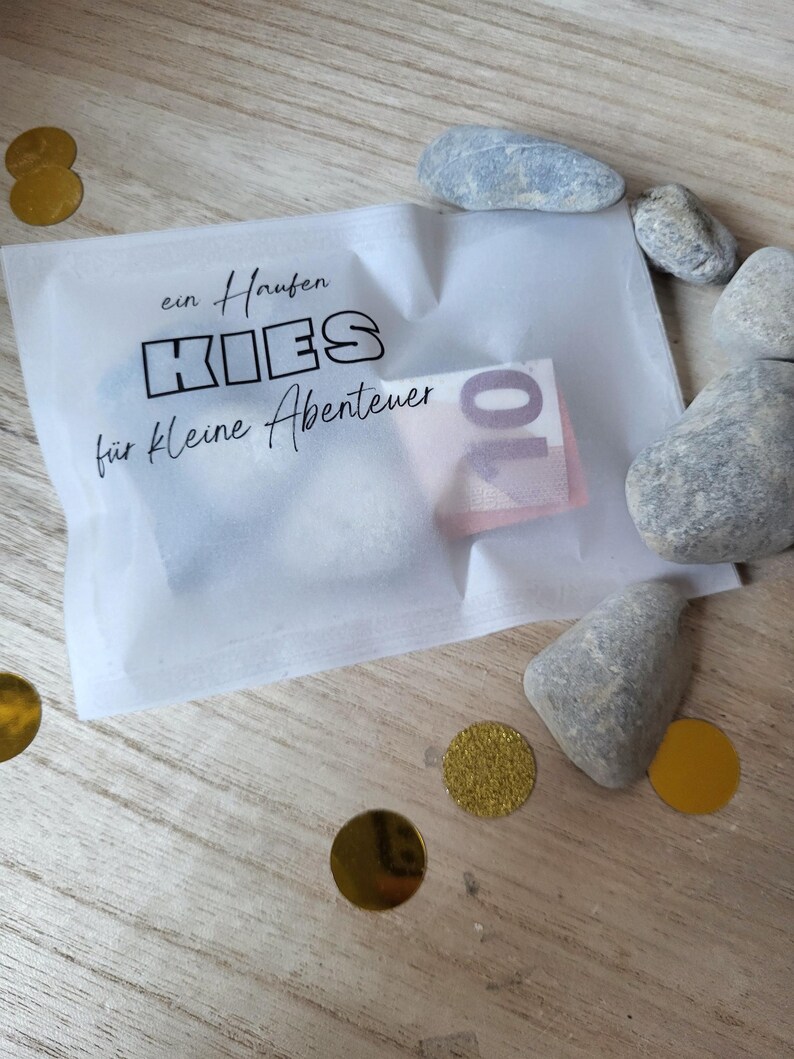 May include: A white translucent paper envelope with the text "ein Haufen KIES f&uuml;r kleine Abenteuer" and a ten-euro note inside. The envelope is surrounded by small grey stones and gold confetti on a wooden surface.