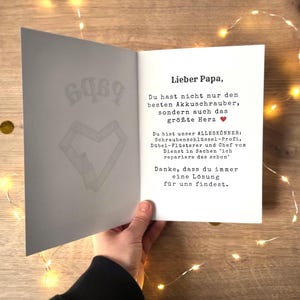 May include: An open greeting card with the text "Lieber Papa" in black font. The card's interior features a message and a heart symbol. The card is held by a person, with a wooden background and string lights.