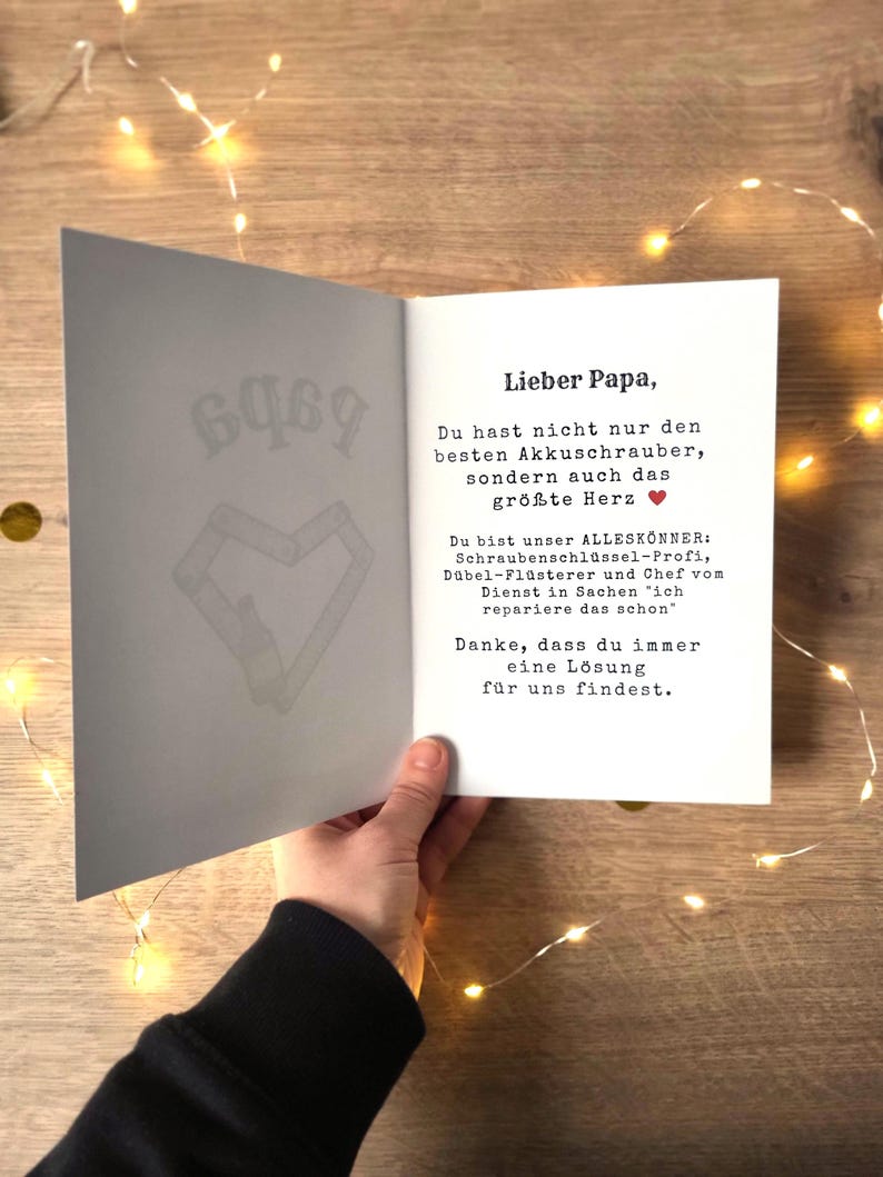 May include: An open greeting card with the text "Lieber Papa" in black font. The card's interior features a message and a heart symbol. The card is held by a person, with a wooden background and string lights.