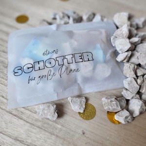 May include: A translucent bag with the German text "SCHOTTER f&uuml;r gro&szlig;e Pl&auml;ne" is surrounded by small, light-coloured rocks and gold confetti. The bag contains a light blue object. The items are arranged on a wooden surface.