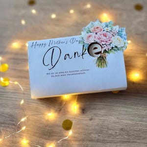 May include: A white Mother's Day card with the word "Danke" and a floral bouquet illustration. The card has the text "Happy Mother's Day!" and a German message. The card is decorated with fairy lights and gold confetti.