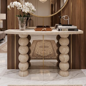 Peut inclure: Un miroir rond à cadre doré fixé sur un mur lambrissé en bois au-dessus d'une console en pierre beige. La table présente deux pieds colonnes à sphères empilées et supporte un vase en verre avec des orchidées blanches, un plateau et des livres.
