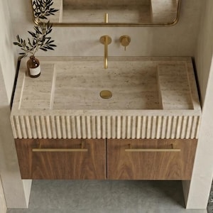 Può includere: Un mobile bagno a parete con lavabo in pietra beige dal frontale scanalato, armadietti in legno scuro con maniglie dorate e un rubinetto dorato. Sopra il lavabo c'è uno specchio ad arco con cornice dorata e una piccola bottiglia di vetro.