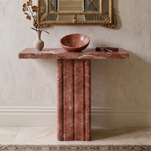 Peut inclure: Une console en marbre rose dotée d'un plateau rectangulaire et d'un pied central cannelé. Sur la table sont posés un petit vase crème avec une tige d'orchidée artificielle, un bol en marbre rose assorti et un trousseau de clés en métal.