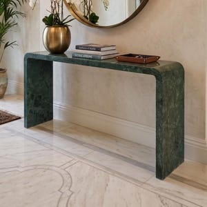 May include: A dark green marble console table with a waterfall edge design. On the table sits a gold metallic vase with white orchids, two stacked books titled Aesthetics of Marble and Global Atriums, and a small brown leather tray.