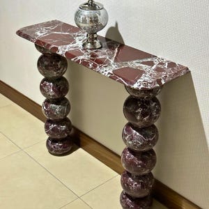 May include: A console table crafted from deep red marble with white veining, featuring a rectangular top and two legs composed of stacked spherical marble spheres. A silver-toned, reflective, mosaic-tiled decorative jar sits on the table.
