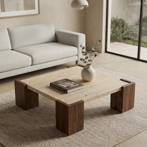 May include: A square coffee table featuring a light beige travertine stone top with a natural porous texture, supported by four thick, dark brown wooden block legs. A small white ceramic vase with dried stems and a book rest on the table surface.