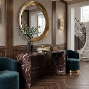Könnte beinhalten: Ein elegantes Interieur mit einem goldgerahmten Spiegel, einem Marmor-Konsolentisch und zwei teal-farbenen Samtsesseln. Der Raum verfügt über dunkle Holzvertäfelungen, einen Kristall-Kronleuchter und einen Fischgrät-Holzboden. Eine schwarze Vase hält Grün.