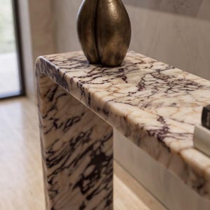 May include: A waterfall console table crafted from white marble with prominent dark purple and black veining. A tall, textured bronze-toned vase with a rounded, bulbous shape sits on the surface, alongside a portion of a book on the right side.