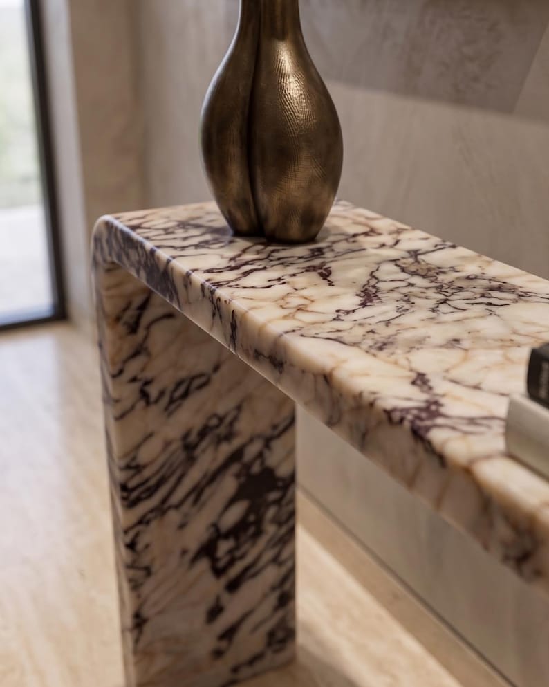 May include: A waterfall console table crafted from white marble with prominent dark purple and black veining. A tall, textured bronze-toned vase with a rounded, bulbous shape sits on the surface, alongside a portion of a book on the right side.