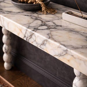 May include: A marble-topped console table with a grey and cream veined pattern. The table has turned legs and is decorated with a dark grey bowl holding prayer beads and a white incense holder. Incense smoke rises from the holder.